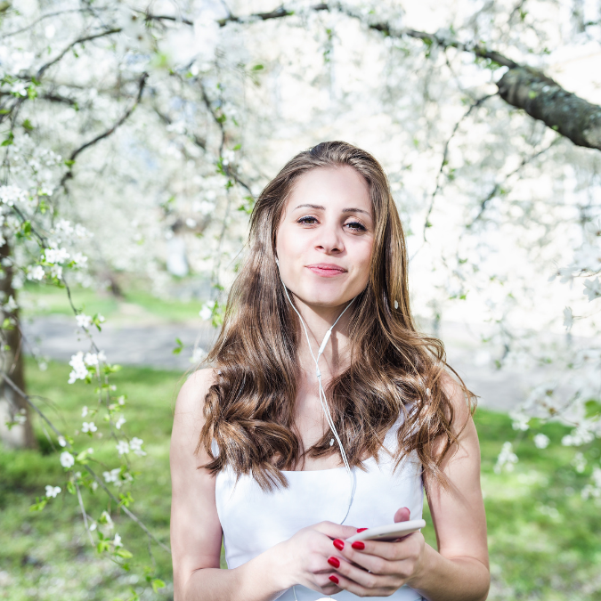 young woman looking pleased while wearing earphones in a park surrounded by white flowers