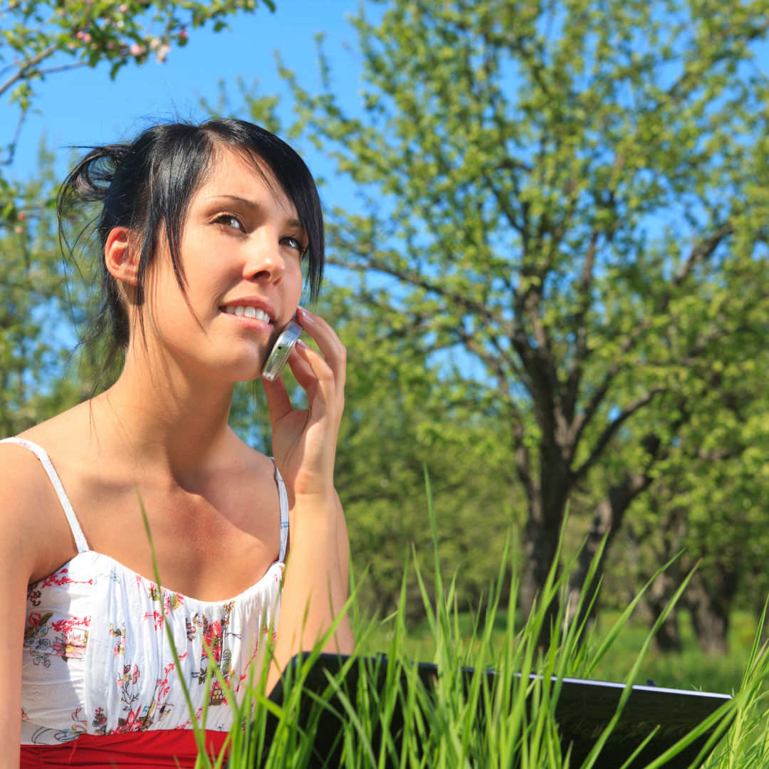 young woman smiling while on her mobile phone sitting on grass in a park