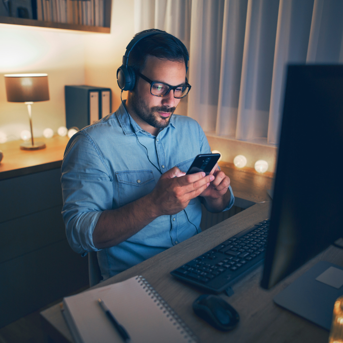 man on his mobile phone wearing headphones sitting at his home office at night time requesting a reschedule of his appointment with Affordable Counselling Solutions